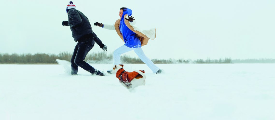 Cheerful couple man and woman running with a dog on a snowy field and having fun. Winter fun with a dog on a walk