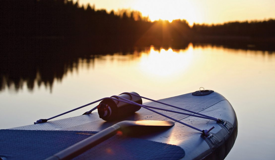 SUP board and surf board with paddle on blue water surface background close up. Surfing and SUP boarding equipment in sunset lights close-up. Outdoor water sports. Surfing lifestyle backgrounds.
